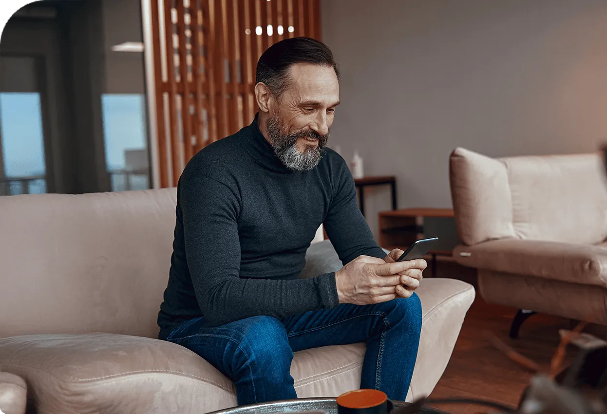 Smiling man sitting on a sofa at home, using a smartphone.
